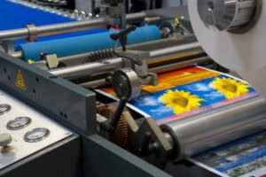 A commercial printing press, equipped with advanced RIP Software, prints colorful sunflower images on sheets of paper, with rollers and control panels visible in the foreground.