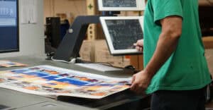 A person in a green shirt checks a color print proof on a table while using RIP Software on a touchscreen monitor in a printing facility.