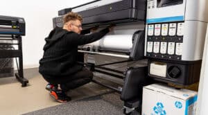 A person is loading a roll of paper into a large-format printer in an office or print shop setting, preparing for printing and cutting tasks using Onyx Graphics software.