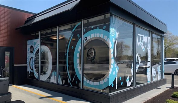 Modern building with large windows decorated with abstract blue, white, and black graphic designs, resembling washing machine doors. Cutting-edge printing by Onyx Graphics enhances the vibrant visuals. Some cars and trees are visible outside.