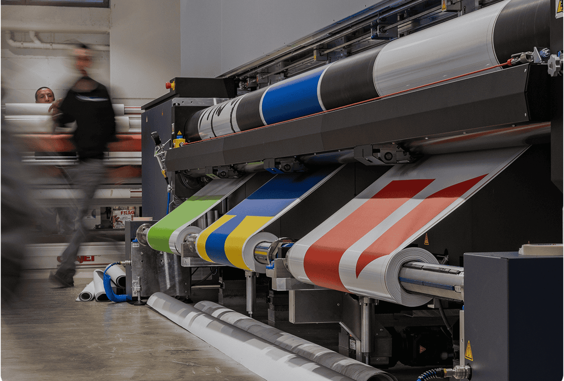 Three large rolls of material in green, blue, and red feed into a printing machine as a person walks by in a print shop, creating the perfect backdrop for a Hero Carousel Test.
