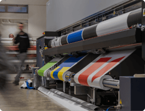 Wide-format digital printer producing colored prints on wide rolls of paper; people are visible in the background of the commercial printing workspace.